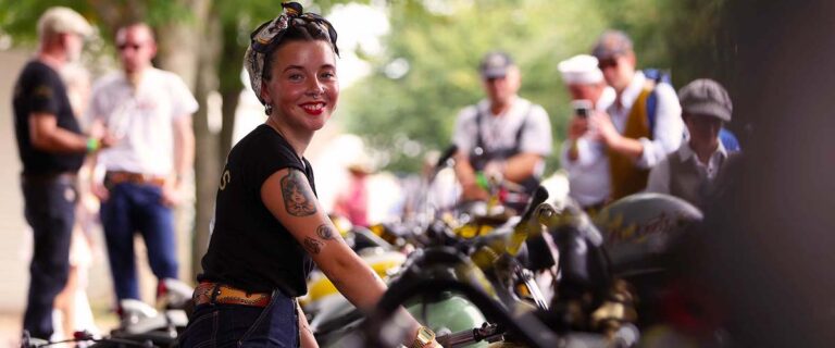 Besucherin im Stil der 1950er-Jahre mit historischem Motorrad beim Goodwood Revival.