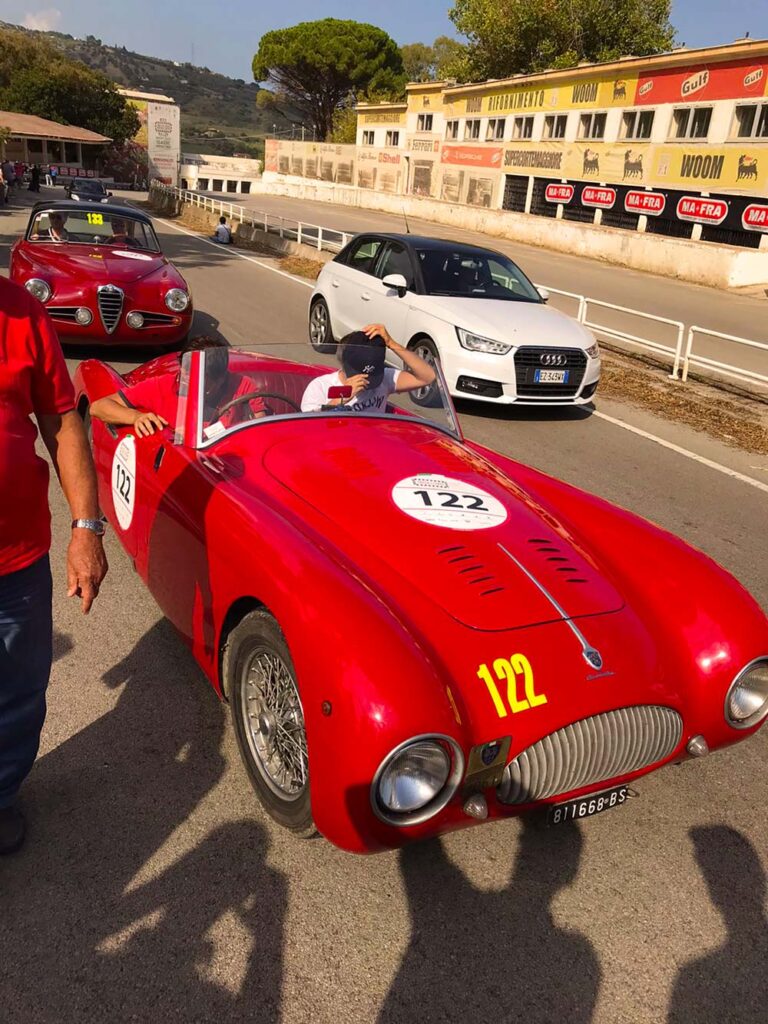 Red Cisitalia 202 Spider at Targa Florio Live in front of the historic Cerda pit buildings in Sicily.